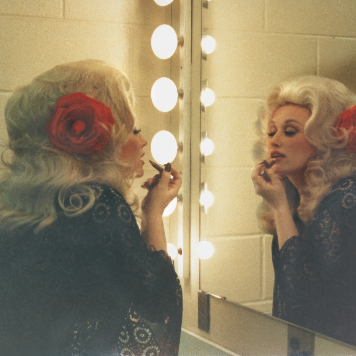 Dolly Parton se maquilla frente al espejo en una imagen que refleja la construcción de su inconfundible estética de pelucas rubias, maquillaje marcado y glamour country. Imagen: archivo