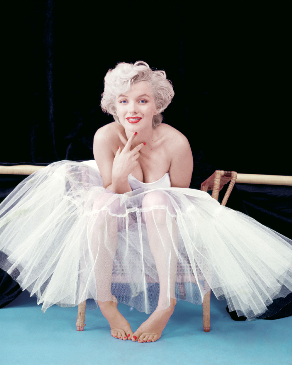 Marilyn Monroe sentada descalza sobre una silla durante una sesión fotográfica, con un vestido blanco de tul, el cabello rubio platino peinado con ondas y los labios rojos. La actriz sonríe a cámara mientras posa con una mano bajo el mentón sobre un fondo negro