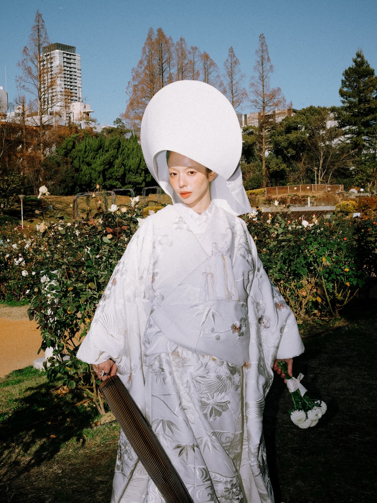 Esta imagen muestra a una novia vistiendo un shiromuku, un kimono nupcial tradicional japonés de color blanco puro adornado con intrincados bordados y complementado con un elegante tocado rígido y redondeado, posando en un jardín al aire libre