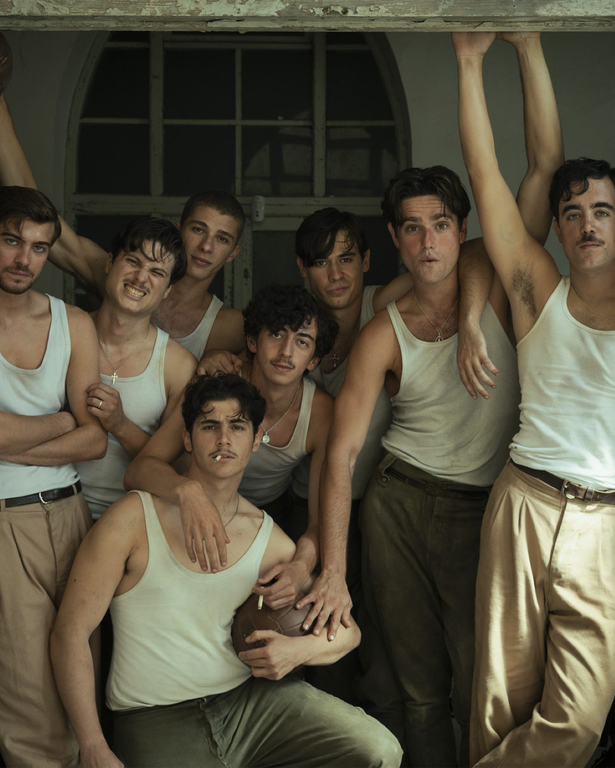 Parte del reparto de «La bola negra» posa junto a una ventana, vestido con camisetas de tirantes blancas y pantalones en tonos neutros, en una de las imágenes promocionales de la película de Los Javis