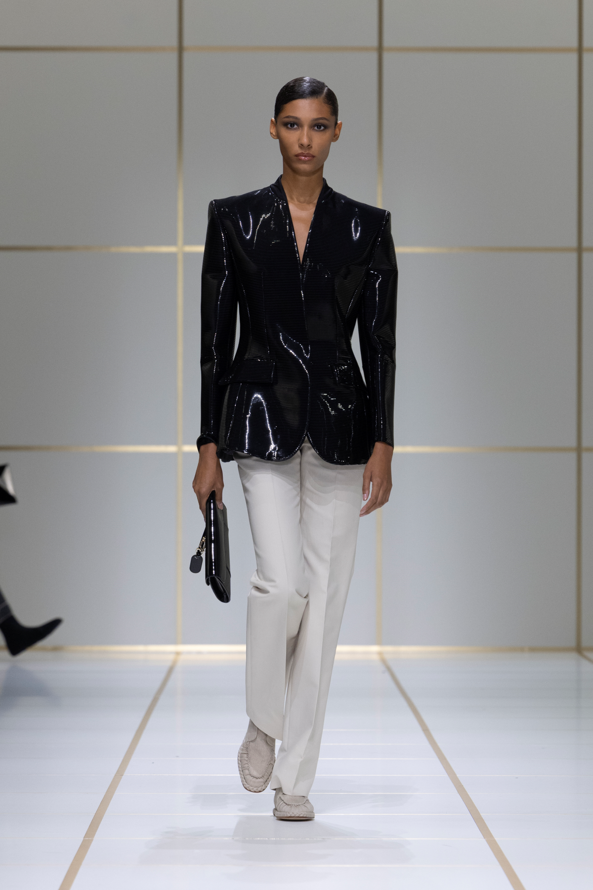 Look 13 de Giorgio Armani Primavera-Verano 2027 «EVOLUTION», con chaqueta negra estructurada y metalizada, hombros en pico, pantalones blancos, bolso pequeño negro de mano y mocasines en tono claro