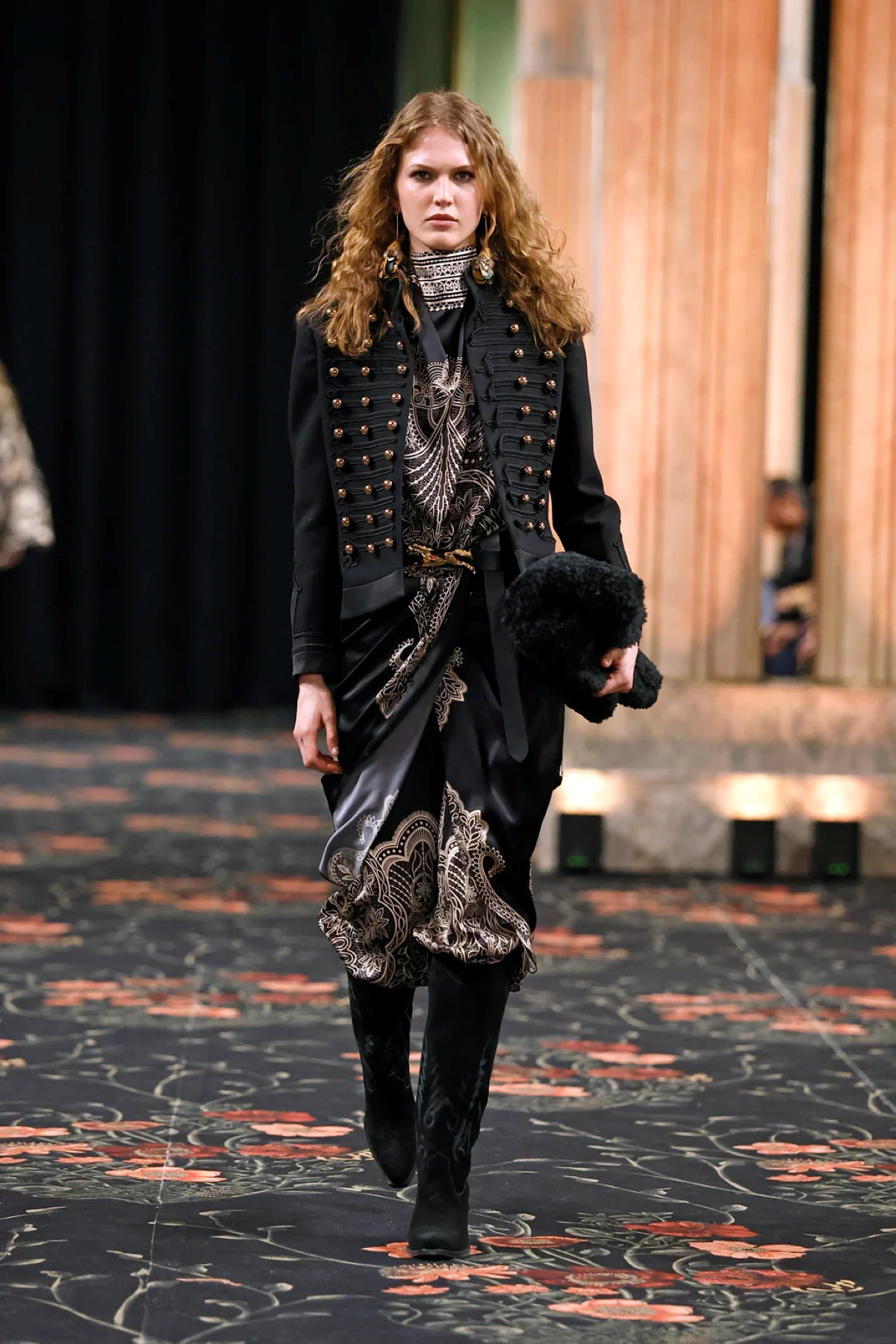 Modelo con chaqueta negra estructurada con botones dorados, vestido estampado y botas altas negras durante el desfile de Johanna Ortiz «Alma» otoño-invierno 2026. Un look de estética ecuestre