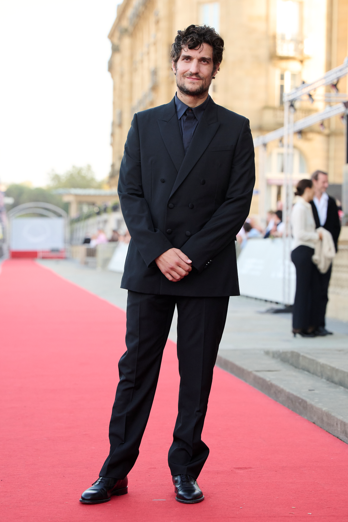 Louis Garrel posa con un traje negro de lana de Dior de doble botonadura y solapas en pico, camisa azul marino y zapatos Derby de piel negra