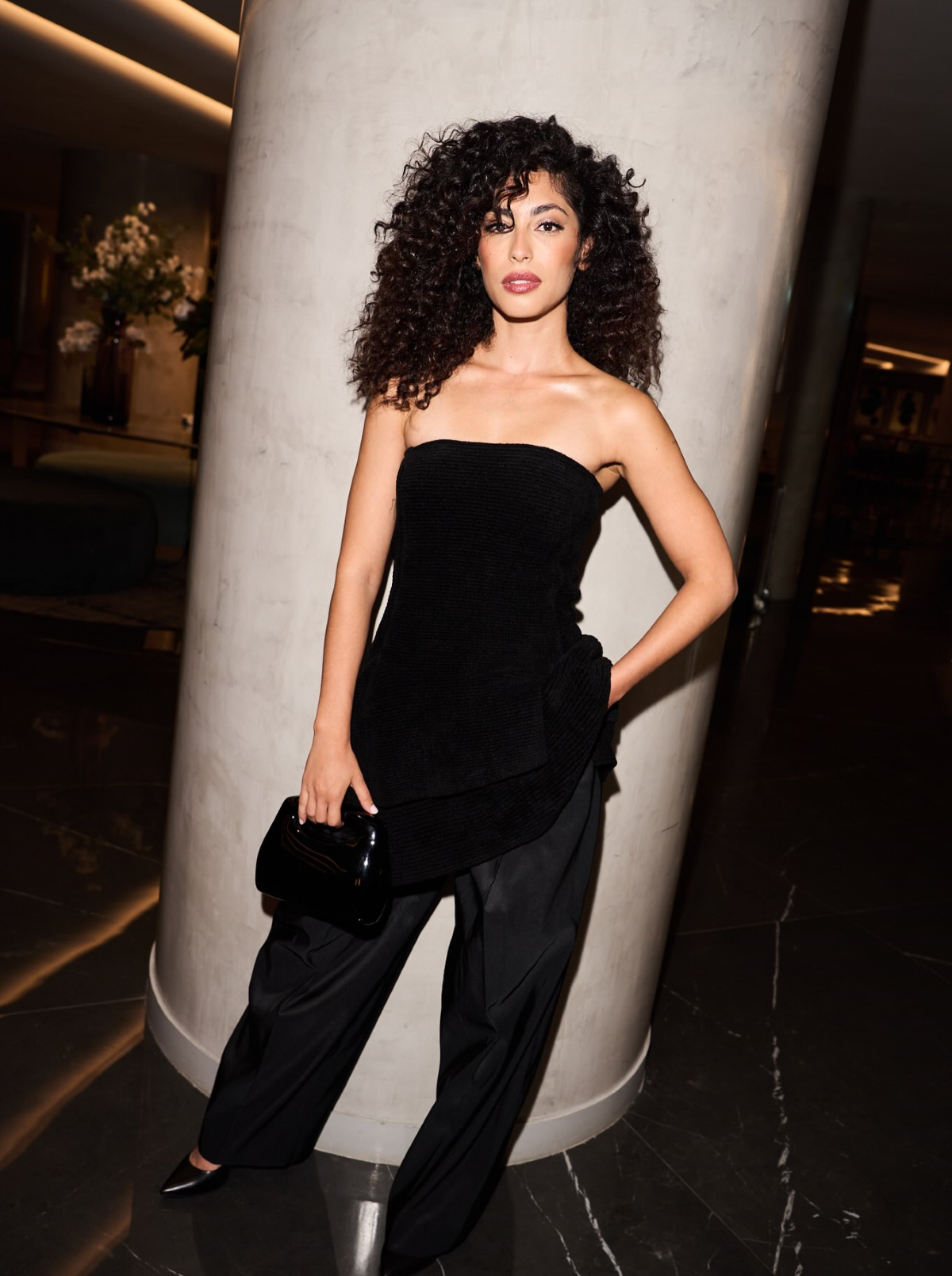 Mina El Hammani posa con un top negro de escote palabra de honor y corte asimétrico, combinado con un pantalón ancho de acabado satinado de Emporio Armani