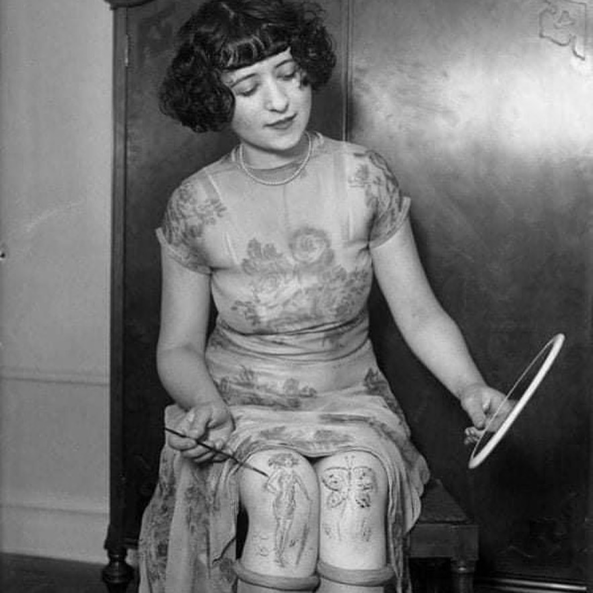 Mujer de los años 20 se pinta una rodilla frente a un espejo para decorar sus piernas desnudas, siguiendo la tendencia de las rodillas pintadas a mano