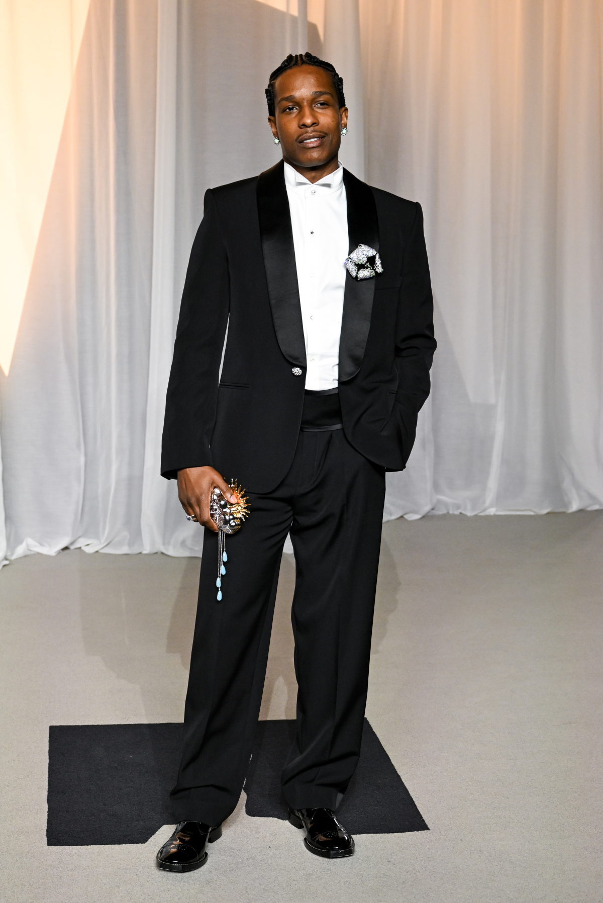 A$AP Rocky con esmoquin negro de CHANEL de textura granulada, cuello chal de satén y botones enjoyados, camisa blanca de piqué con botón de perla, pendientes, broche, bolso y zapatos de la firma 
