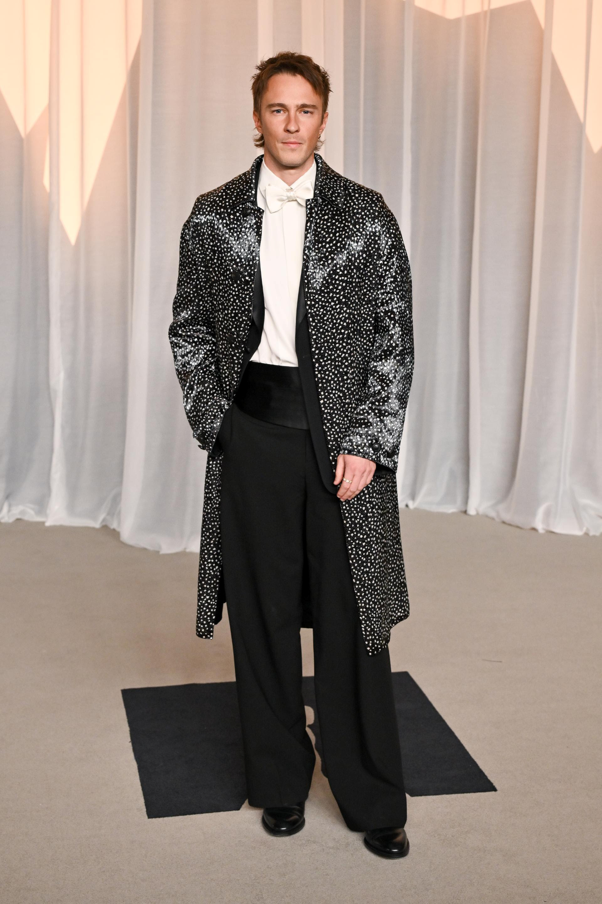 Drew Starkey con abrigo de seda blanco y negro de Dior sobre esmoquin negro de lana y seda, camisa blanca de algodón, pajarita blanca de seda, faja negra y zapatos derby de piel negra en la Academy Museum Gala 2026