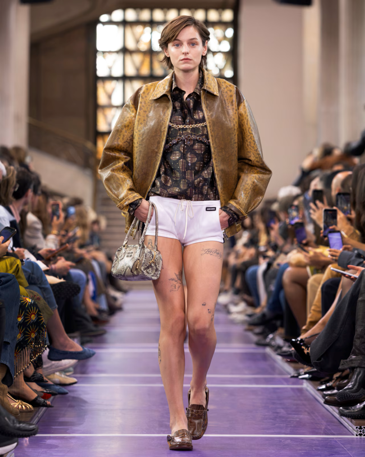 Emma Corrin desfila para Miu Miu Primavera/Verano 2027 con chaqueta de cuero marrón, camisa estampada y microshorts blancos