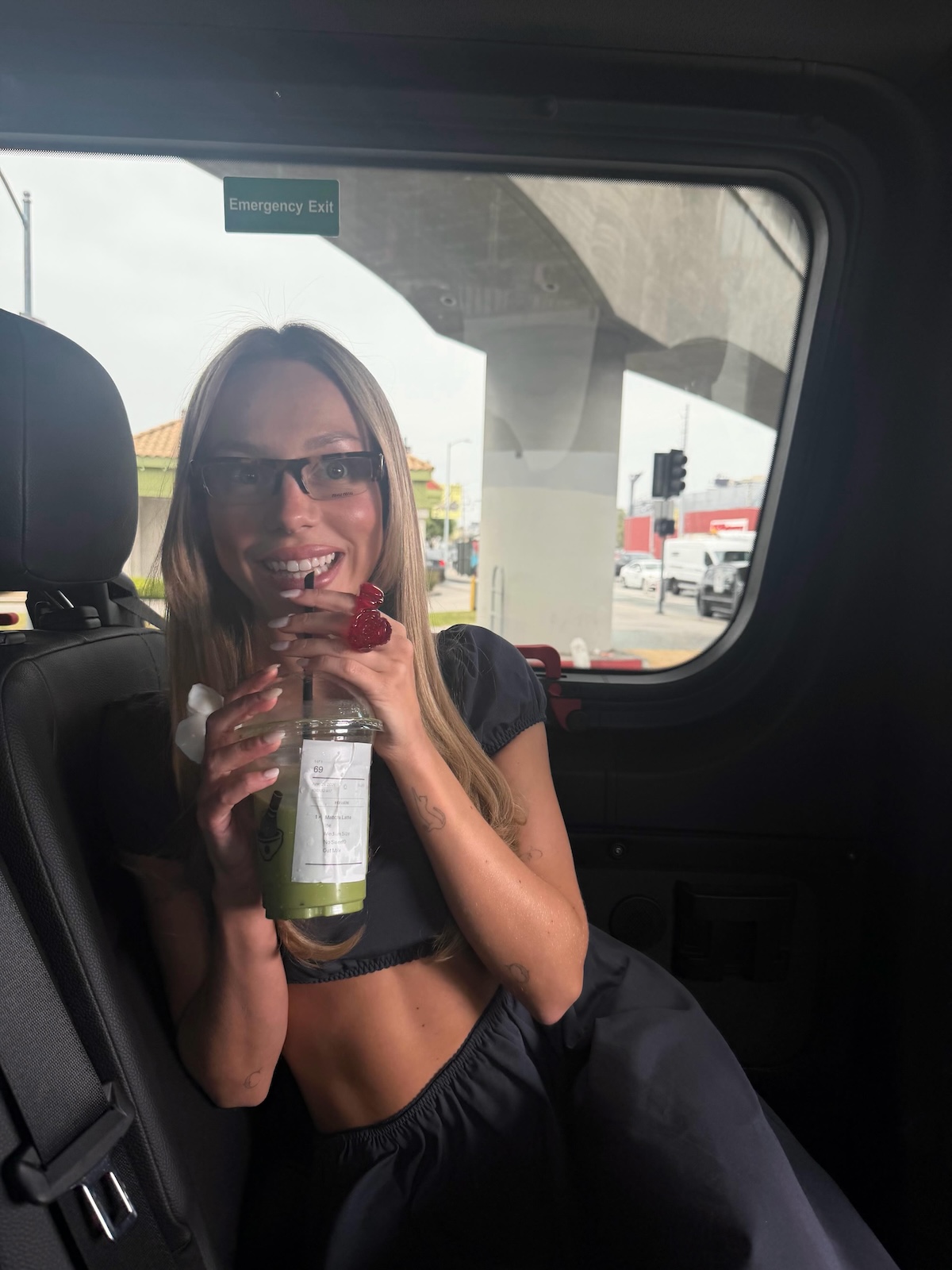 Ester Expósito en el asiento trasero de un vehículo, usando gafas de sol oscuras y bebiendo un matcha con hielo