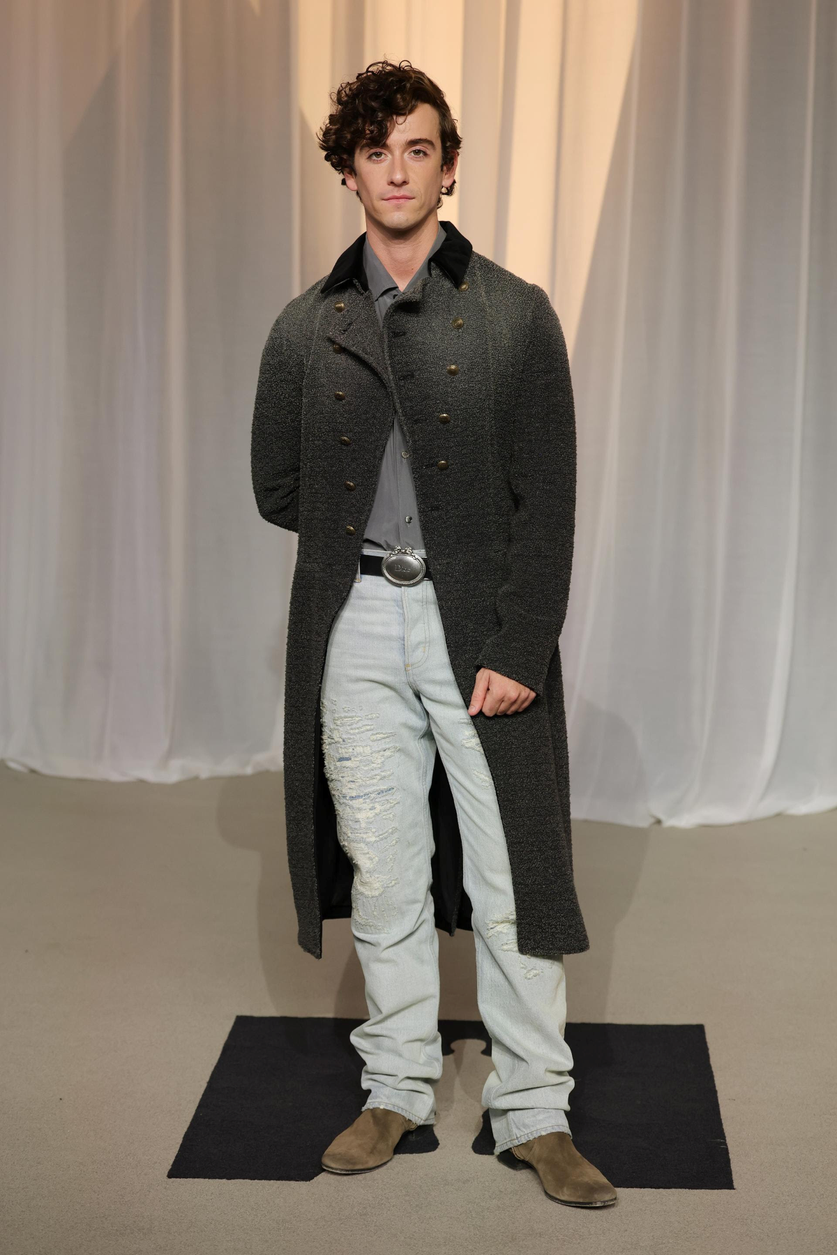 Guitarricadelafuente con chaqueta larga cruzada de lana gris oscuro de Dior, camisa de seda gris, vaqueros azul claro rasgados, cinturón negro de piel con medallón y botas de ante marrón en la Academy Museum Gala 2026.