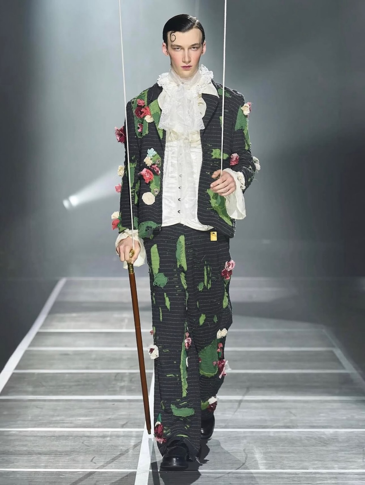 La imagen muestra a un modelo desfilando con un conjunto de chaqueta y pantalón estampados con flores y detalles de encaje de la colección Primavera/Verano 2025 de la marca KidSuper, diseñada por Colm Dillane. Imagen: @enigma.curation
