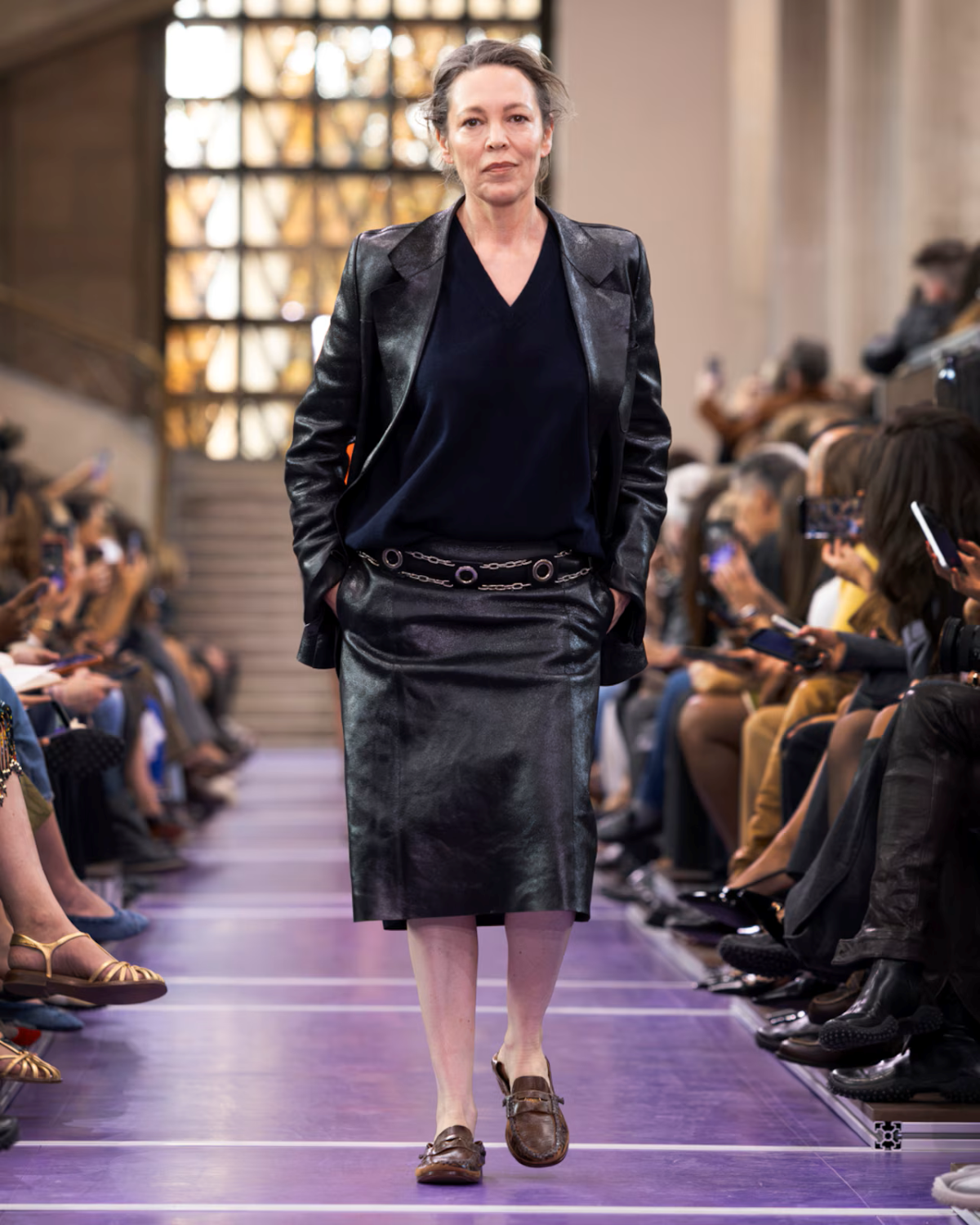 Olivia Colman desfila para Miu Miu Primavera/Verano 2027 con blazer y falda de cuero negro, jersey azul marino y mocasines marrones